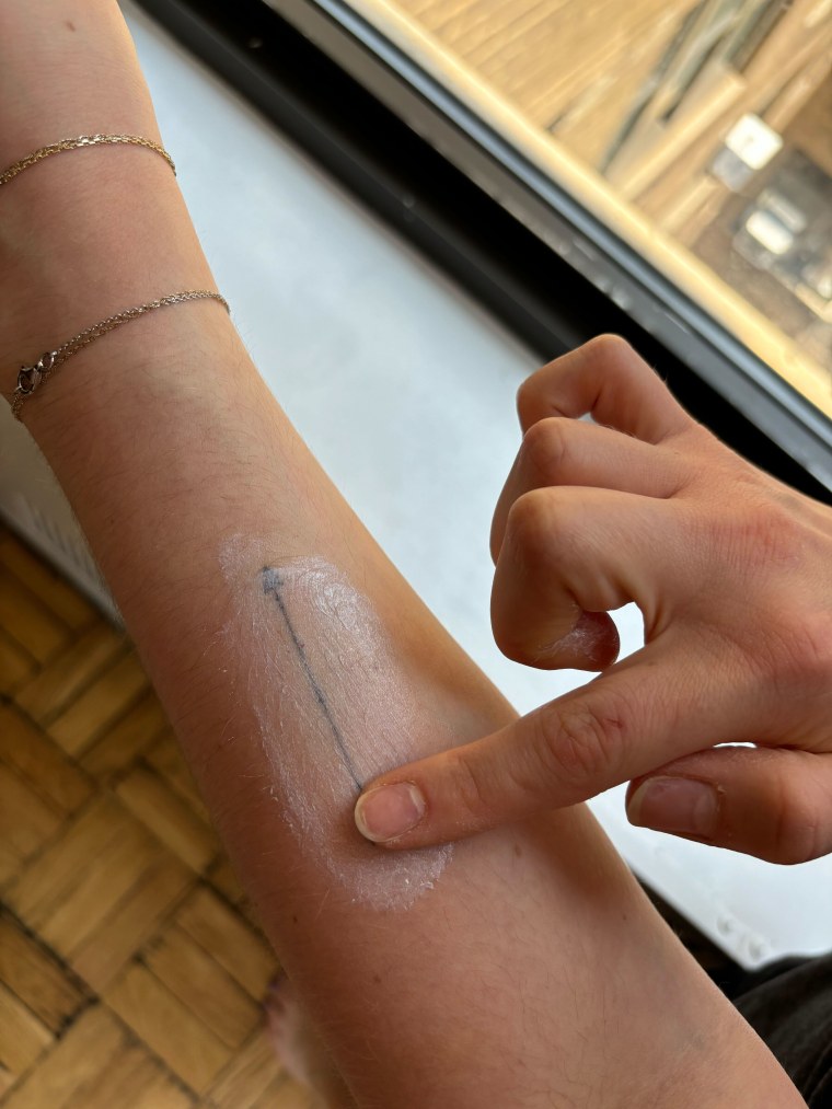 A finger applying white sunscreen to a forearm tattoo of an arrow.