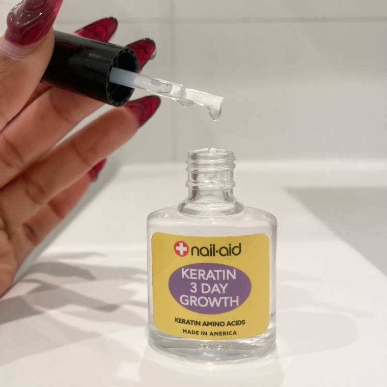 The Nail-Aid Keratin treatment on a white bathroom counter against a white wall, with the applicator held above the bottle by a left hand with red nails.