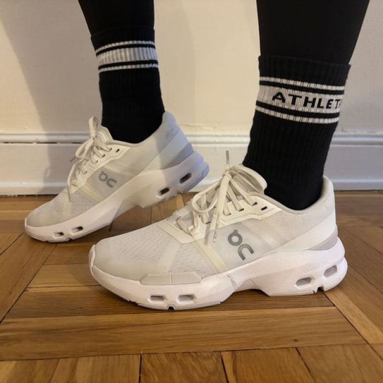 A woman wearing a pair of On Cloudpulse training sneakers while standing on a wood floor in front of a white wall.