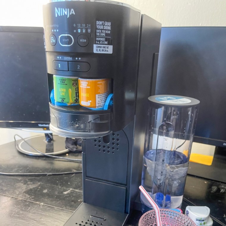 Ninja Thirsti Drink System Soda Maker sitting on a countertop beside a glass with a straw in it.