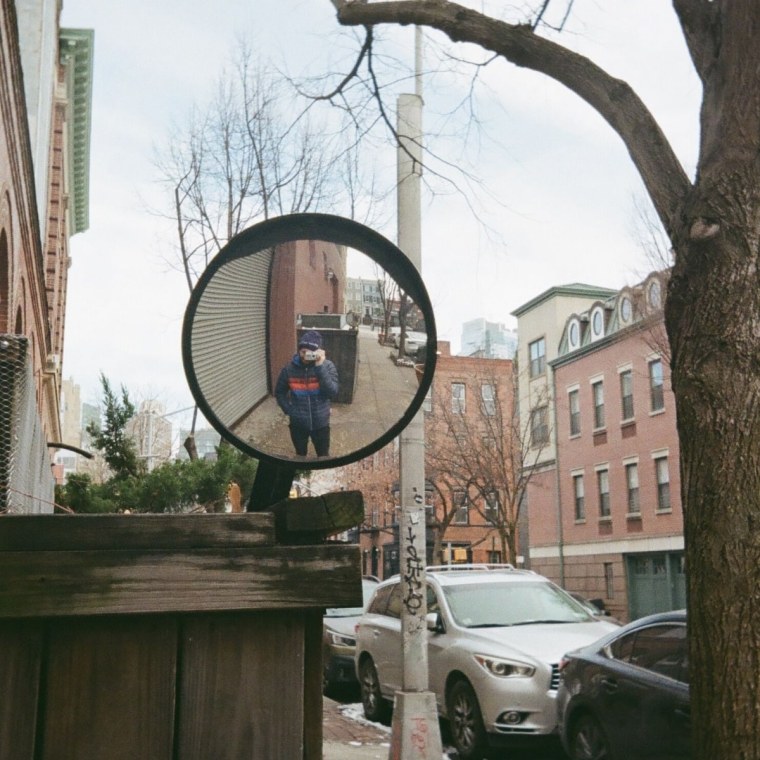 A photo of a driveway mirror on a sidewalk, with the photographer in the mirror’s reflection.