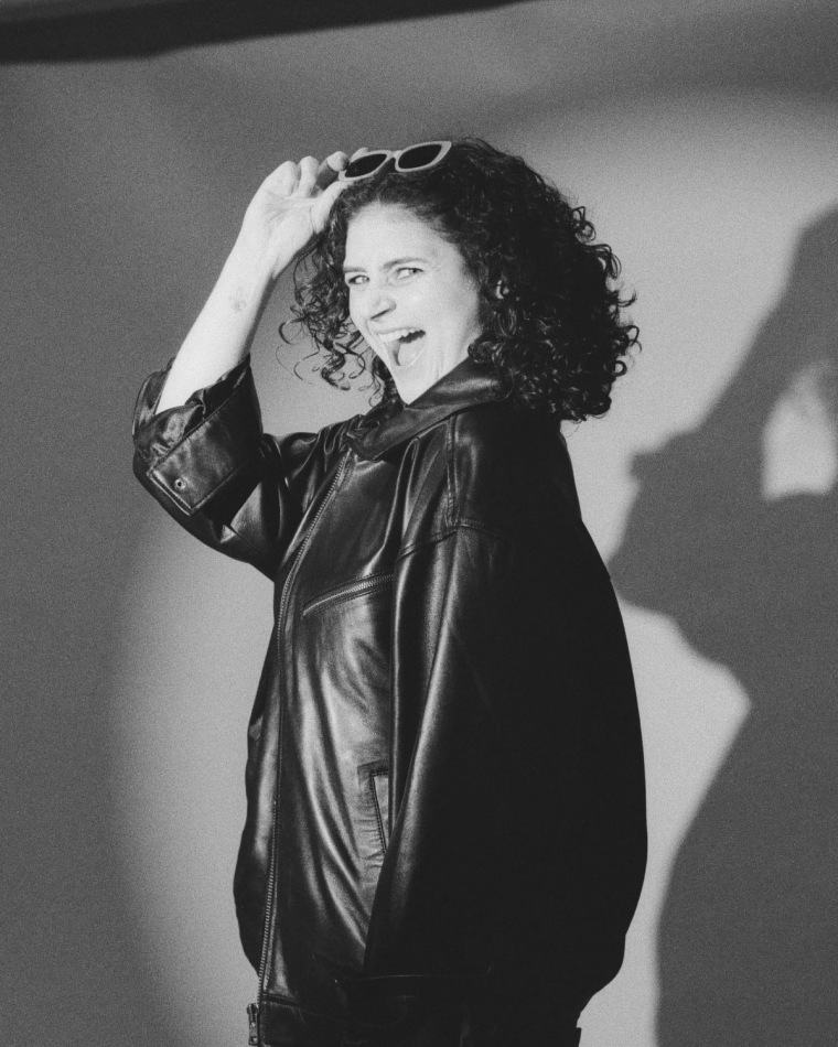 A black-and-white photo of a woman in a spotlight against a white background, smiling over her shoulder at the camera.
