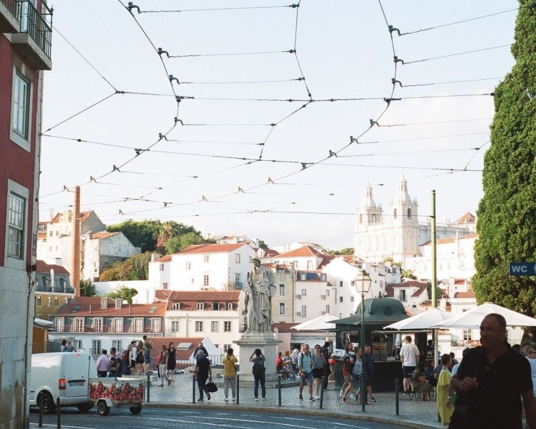 A layered city street with tourists photographing an old statue in front of bright white and brown buildings.