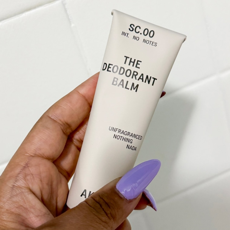 The AKT Deodorant Balm held up by a left hand against a white tile background.