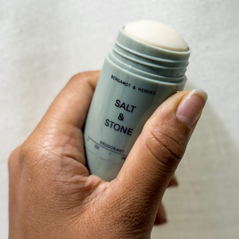 Salt and Stone Natural Deodorant stick with the cap off, held up by a left hand against a white background.